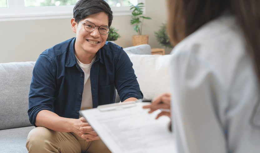 A man sits on a couch smiling and talking to a person with a clipboard.