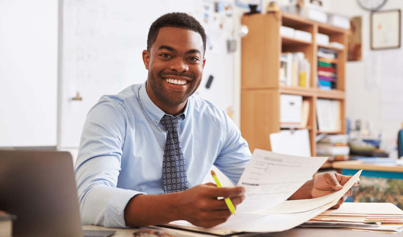 A man in a classroom seated at a desk, smiling while holding papers and a highlighter.