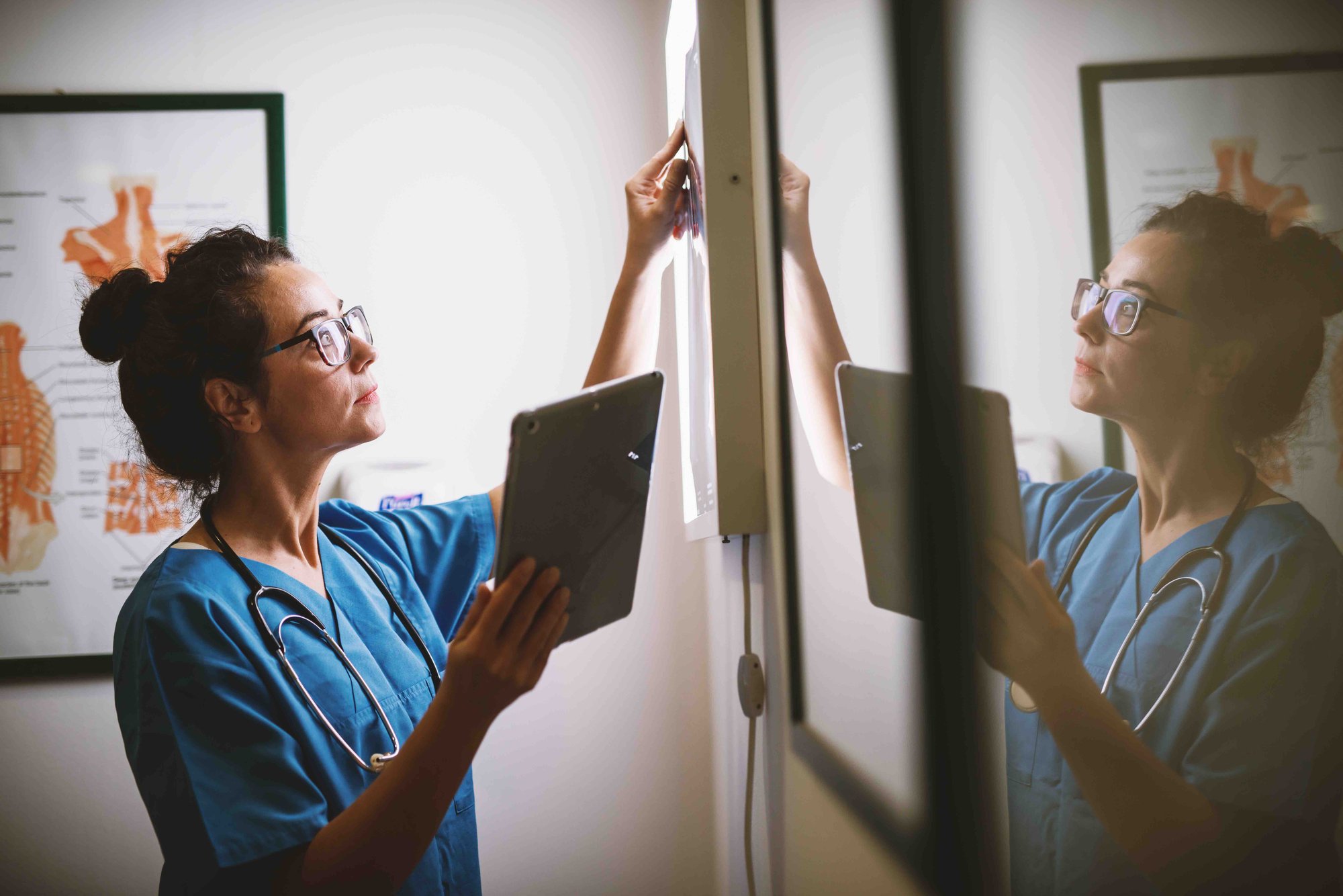 A female medical professional in scrubs and glasses adjusts a spinal x-ray on a lightbox while holding a clipboard.