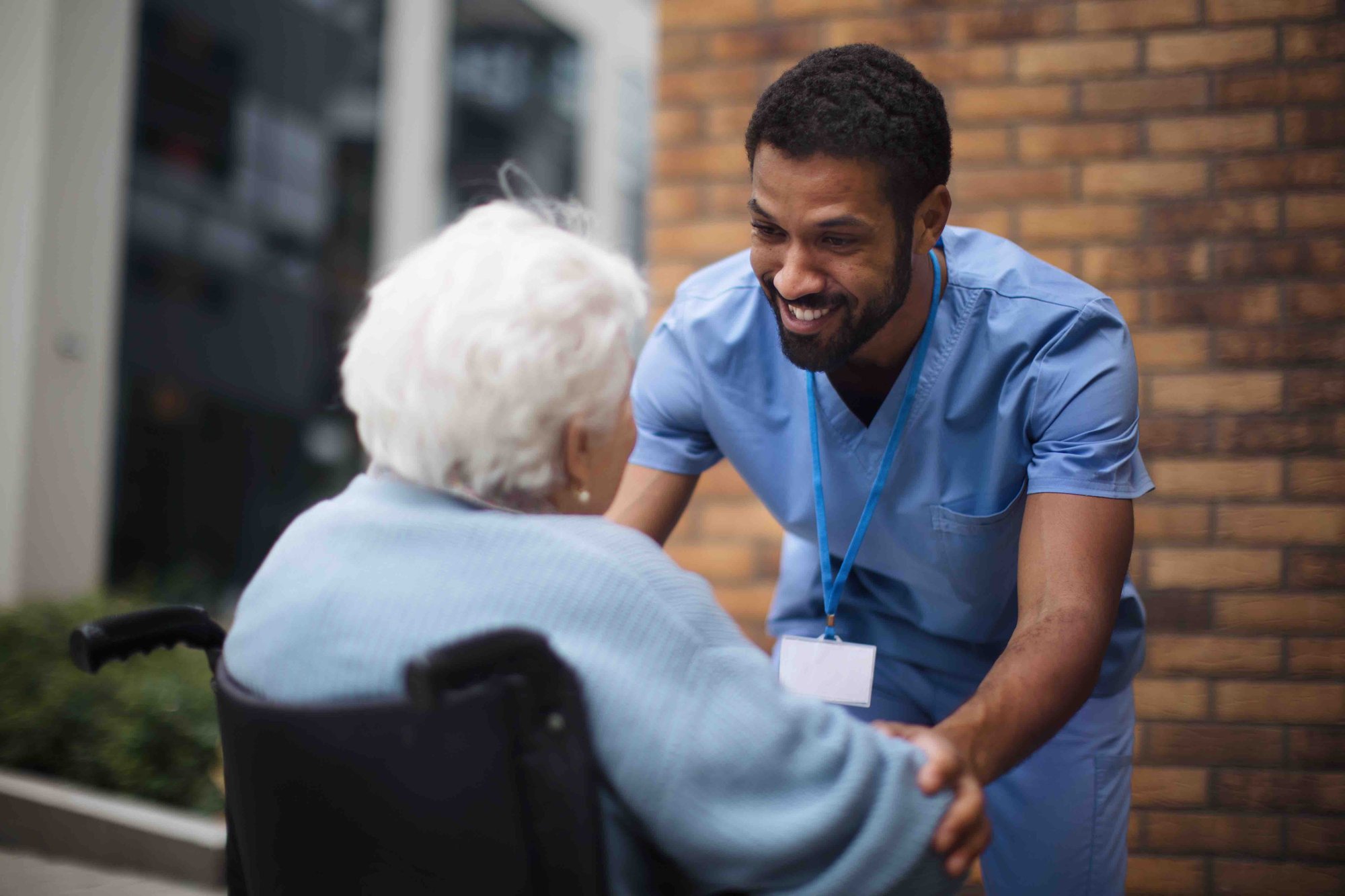 A male nurse in blue scrubs smiles at an elderly woman in a wheelchair, holding her hands outside a building.