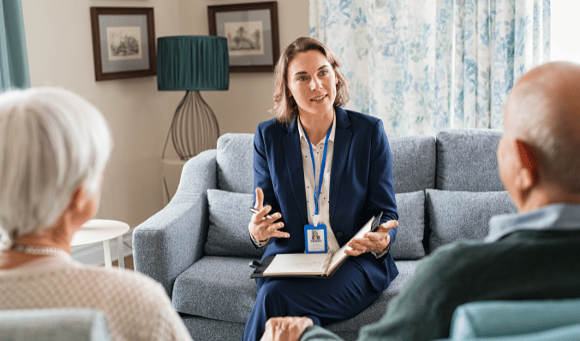 A female social worker, holding a notebook and speaking to an elderly couple seated across from her in a well-lit living room.