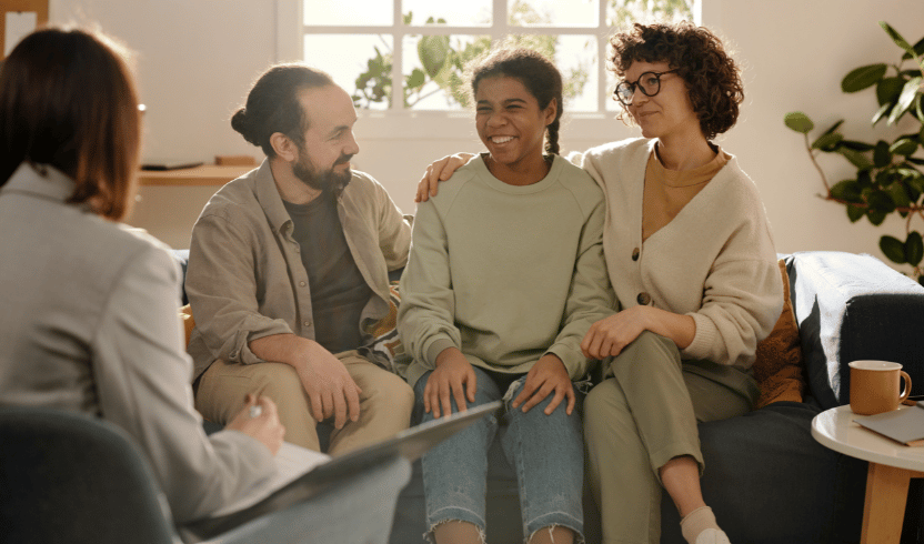 A smiling teenager sits between two supportive adults on a couch, while another person takes notes in the foreground.