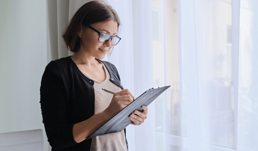 A woman wearing glasses stands by a window holding a pen and writing on a clipboard. She is dressed in a black cardigan over a beige top.