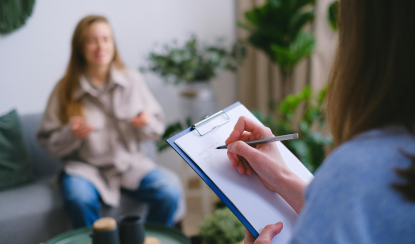 A person holding a clipboard and pen is taking notes while another person sits in the background, gesturing with their hands.
