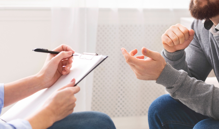 Two people seated, one holding a clipboard and pen, appearing to be in a discussion.
